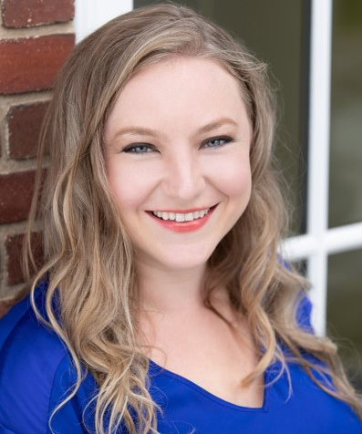 Amanda Schmitt smiling at the camera with a brick wall and window behind her.